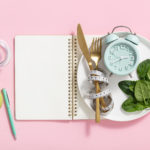 Composition with cutlery, measuring tape, blank paper notebook, spinach, apple, water and alarm clock on color background. Diet concept, copy space